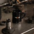 Load image into Gallery viewer, Black mug with a logo on a metal surface with mechanical parts