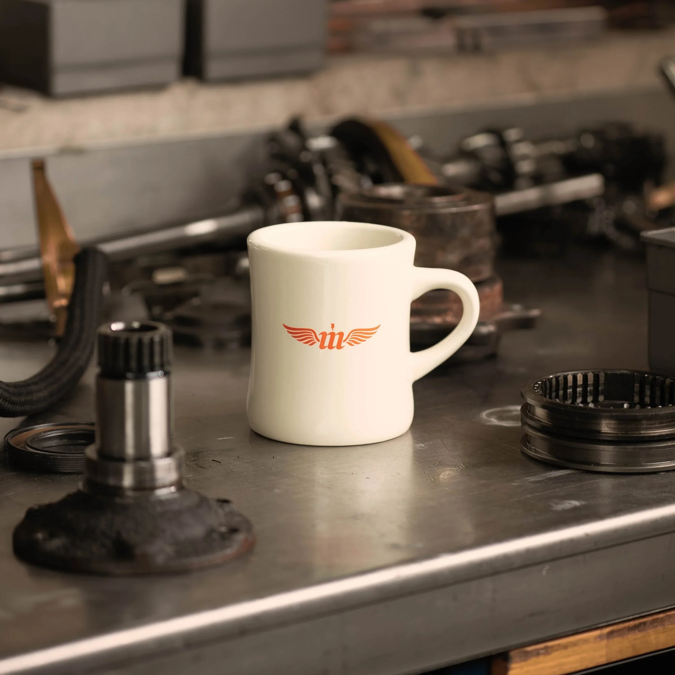 White mug with a logo on a metal workbench with mechanical parts